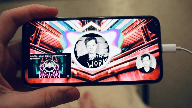 Aluminum framed smartphone displaying a colorful music video playlist on a streaming app with vibrant visuals and artist imag