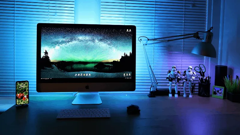 IMac computer with starry night sky wallpaper, surrounded by gaming and tech accessories on a modern desk illuminated by blue