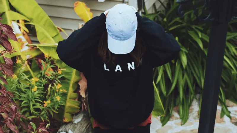 A young woman in a black sweatshirt and light blue cap stands outdoors amid lush tropical plants and greenery, capturing a re