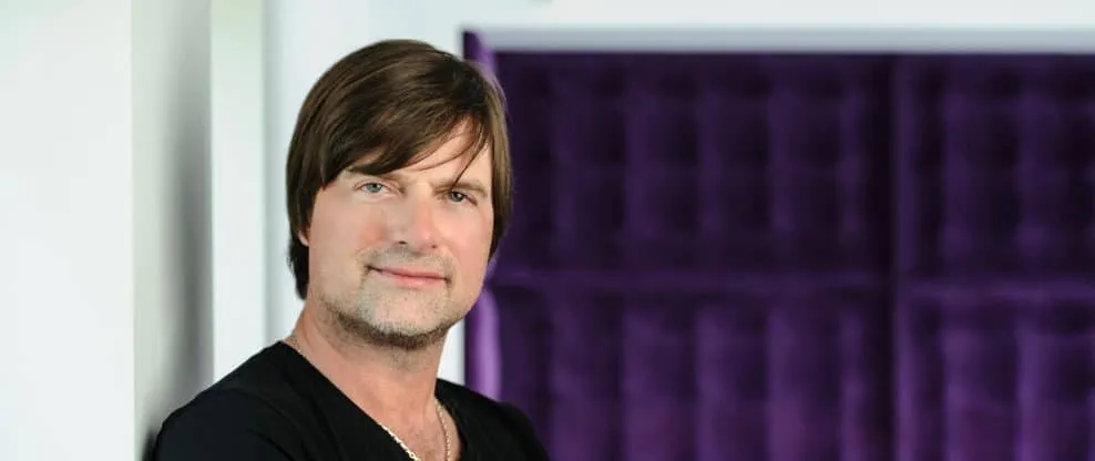 Professional male artist with dark brown hair and a slight smile, standing in front of a purple soundproof background, repres