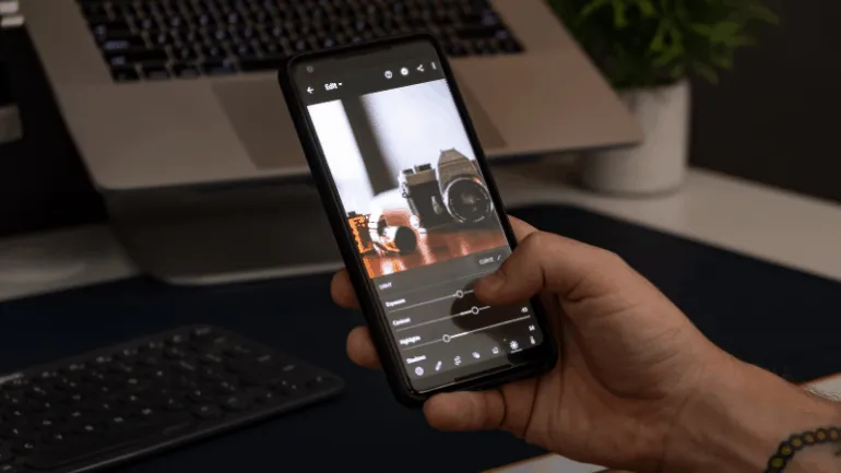 A person editing a photo of a vintage camera on a smartphone in front of a laptop and keyboard, showcasing digital content cr