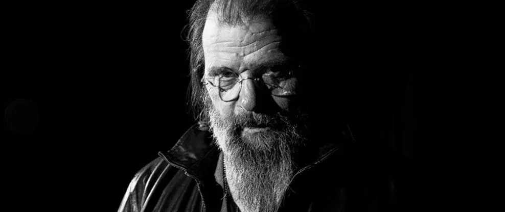 Old man with glasses and beard in black leather jacket, dark background, artistic portrait, music industry expert, Hypebot so