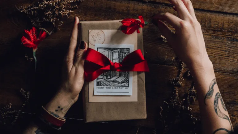 Hand wrapping a gift with a red ribbon on a rustic wooden surface, surrounded by decorative paper shreds, highlighting holida