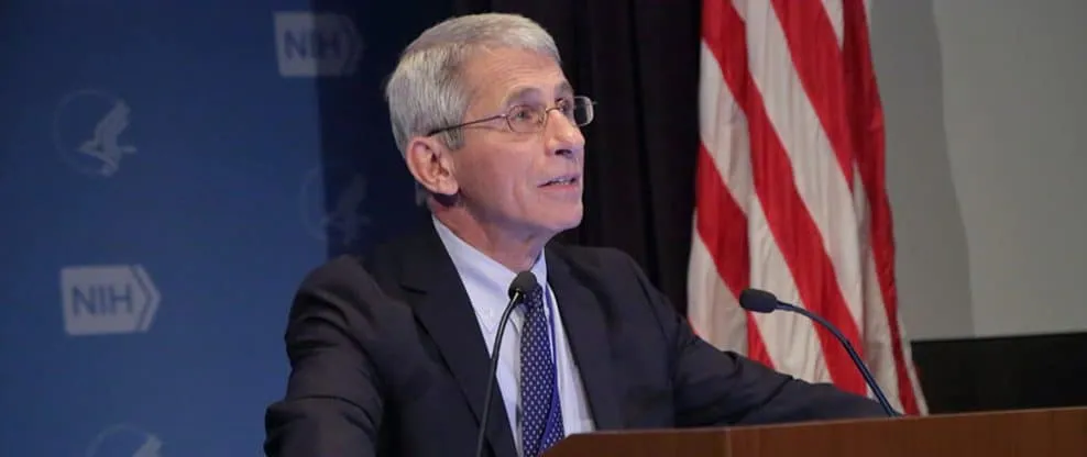 Senior male speaker at a press conference or panel discussion, with an American flag and NH logo in the background, deliverin