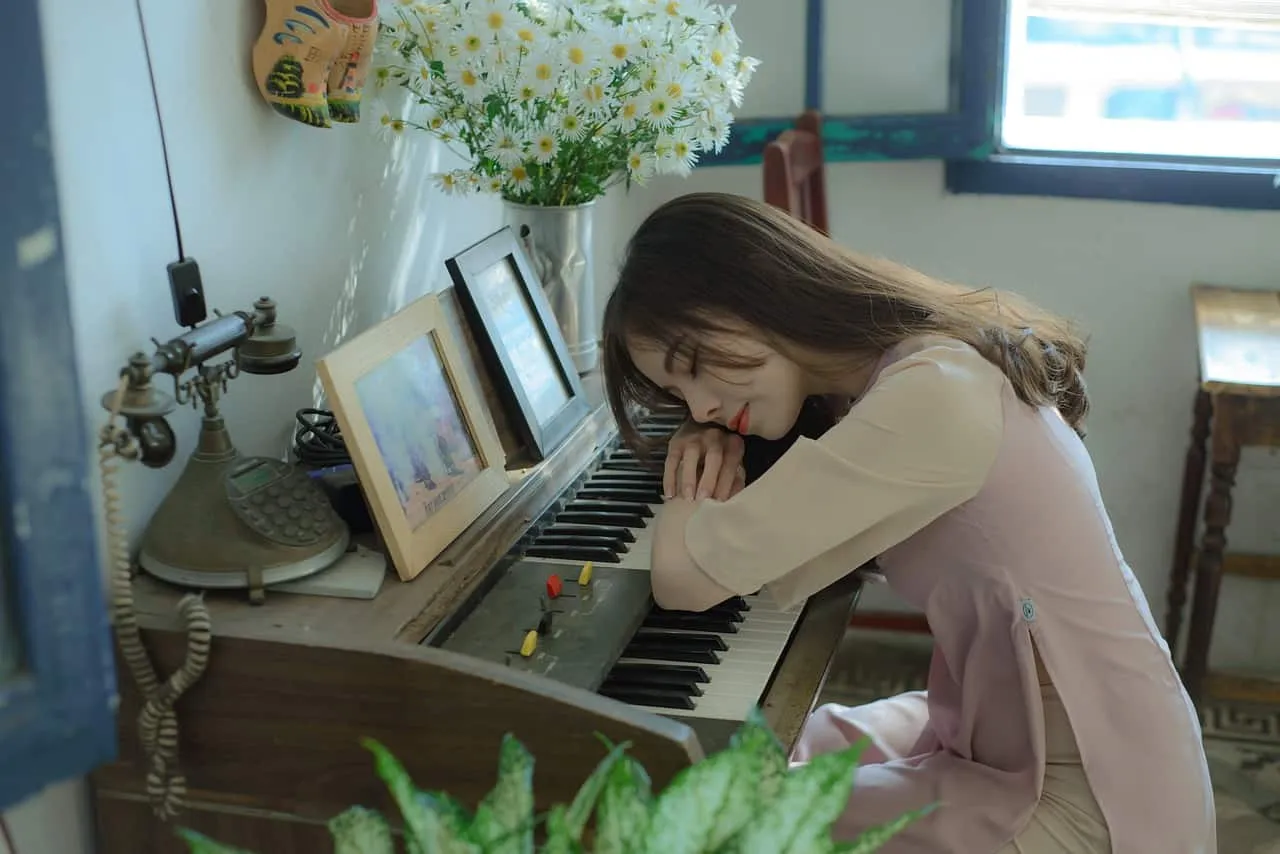 Soft pink dress wearing woman resting her head on a vintage keyboard piano in a cozy, well-lit room with flowers and framed p