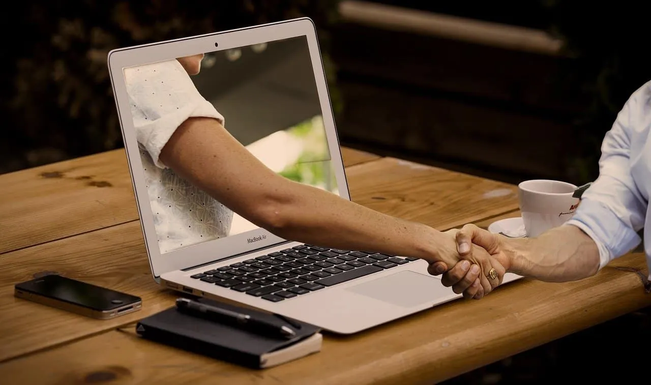 Handshake occurring through a laptop screen between two people working remotely, symbolizing digital collaboration and online
