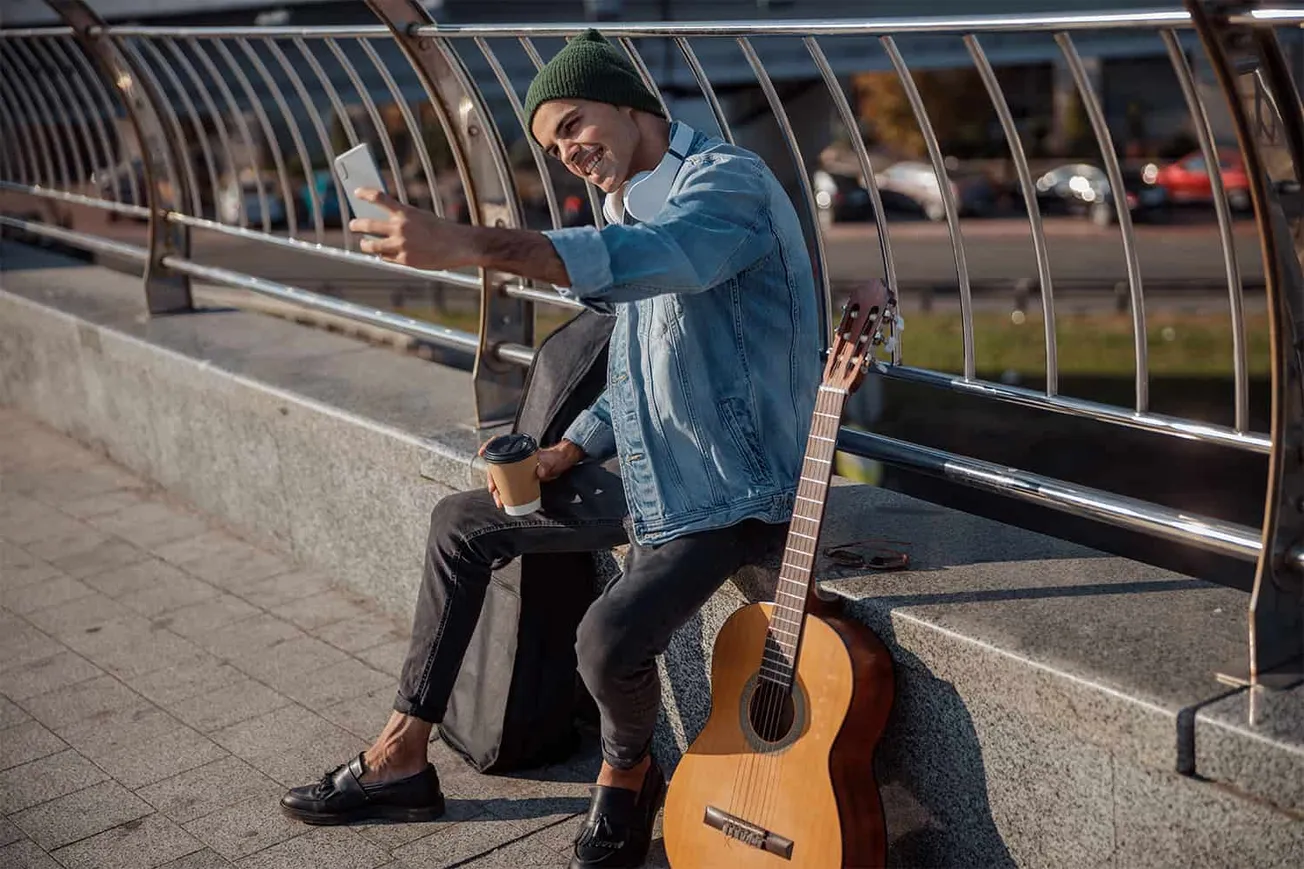 Young man taking a selfie at an urban park, sitting on a bench with a guitar and coffee, in a casual street scene promoting m
