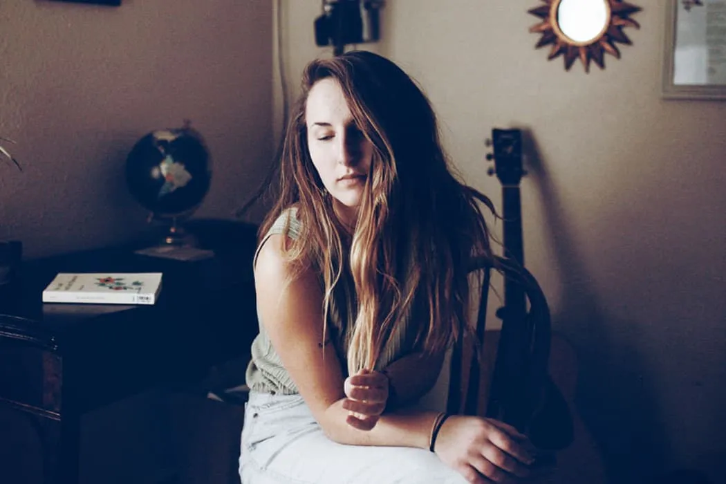 Reflective young woman with long hair sitting on a wooden chair in a cozy room, warm lighting, guitar and globe in the backgr