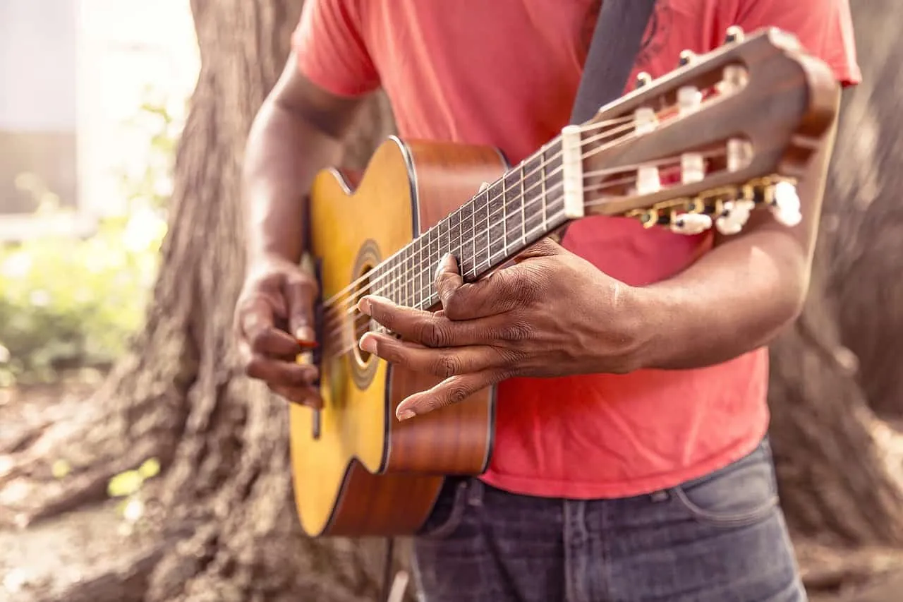 A person playing acoustic guitar outdoors near a large tree, showcasing musical talent and outdoor music recreation.