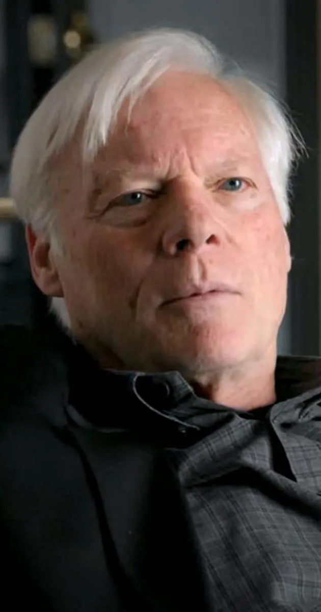 Aging man with silver hair and blue eyes, wearing a dark blazer and plaid shirt, sitting indoors with a serious expression, c