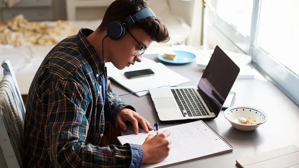 Studious young man in headphones studying at a desk with laptop, notebook, and snacks, creating a focused environment for onl