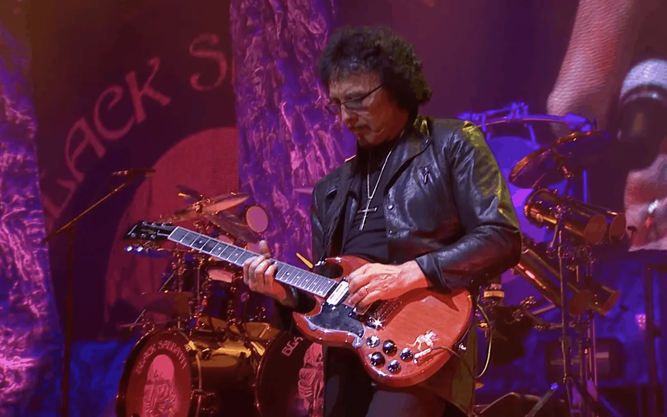 Guitarist performing live on stage with drum set in background, colorful lighting, and rock music atmosphere at a concert.