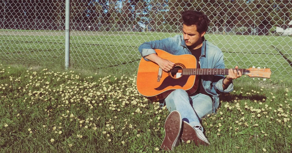 Guitarist sitting on grass near a chain-link fence, playing acoustic guitar outdoors in a park setting, capturing musical cre