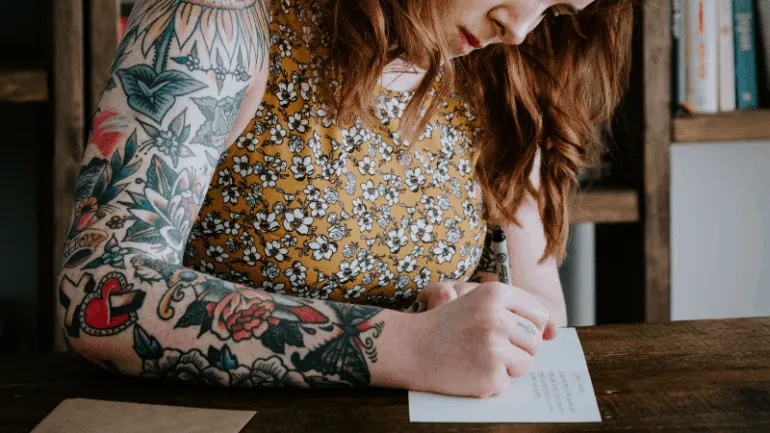 Tattooed woman writing on paper at wooden desk, creative workspace, vibrant tattoos, floral dress, focused, artistic expressi