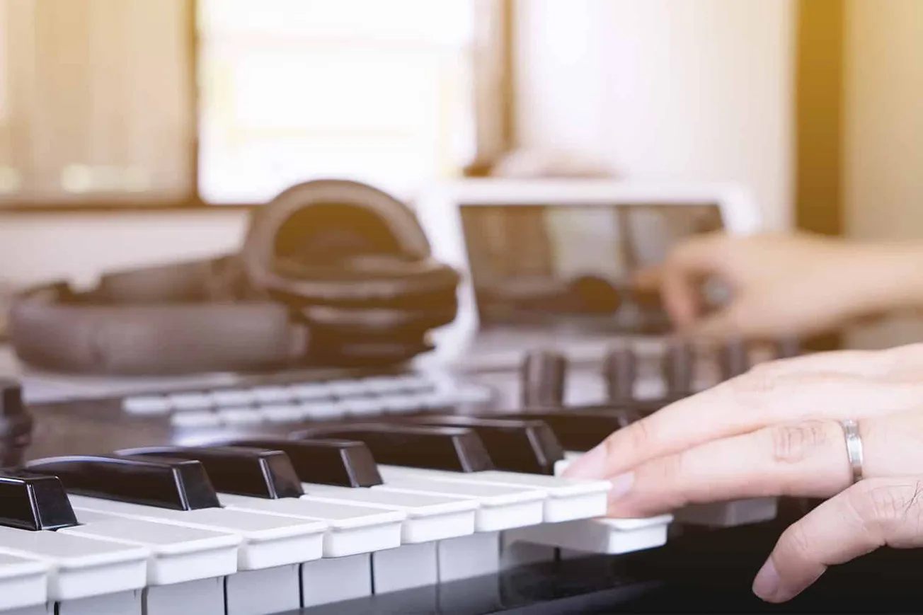 Close-up of a person's hand playing piano keys in a music studio or home setting, with headphones and electronic equipment in