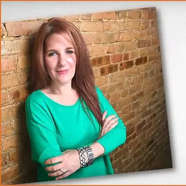 Young woman with red hair wearing a bright teal top, smiling confidently against a rustic brick wall background.
