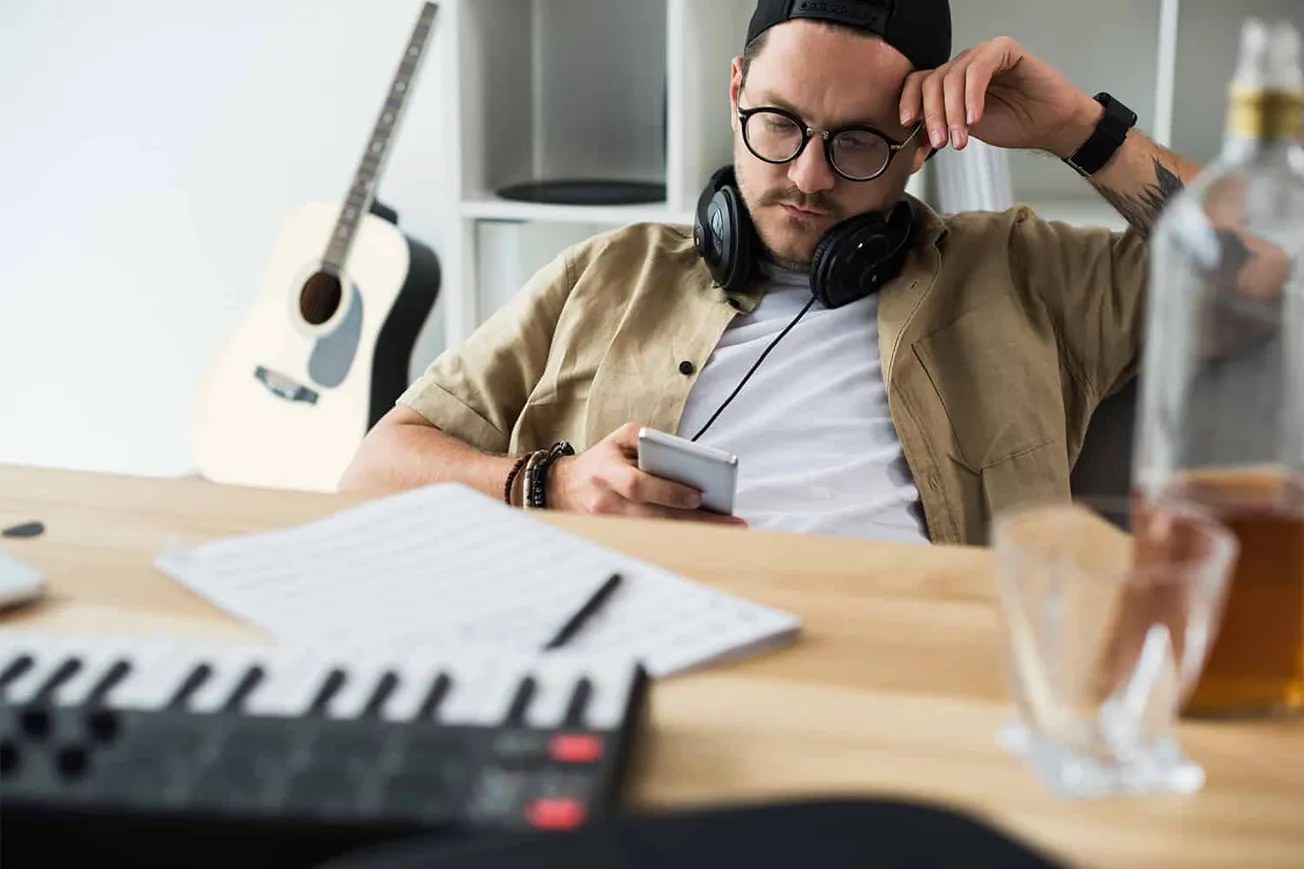 Creative musician using smartphone in home studio with guitar, headphones, mixing console, and music sheets, representing mus