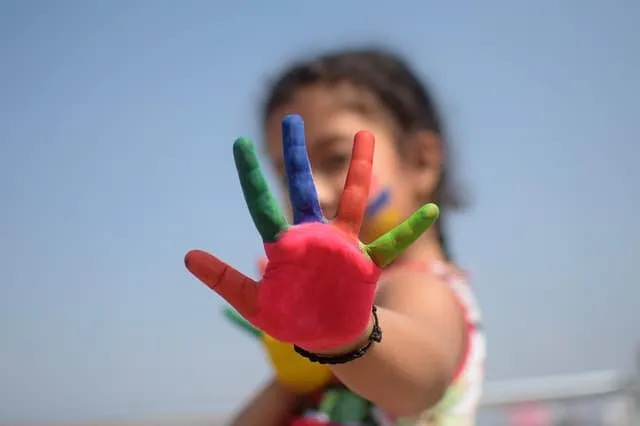 Colorful painted hand reaching out towards the camera with a blurred child background, symbolizing creativity and artistic ex