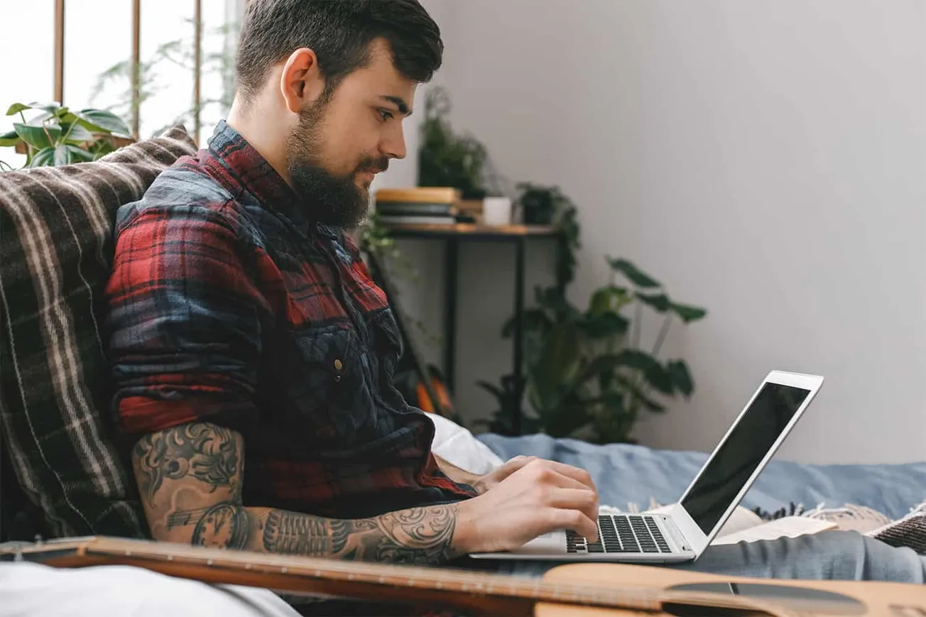 Young man using a laptop in a cozy home office, surrounded by plants, working or browsing online, representing modern digital