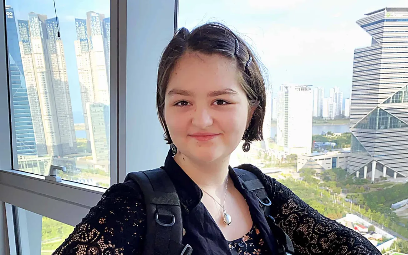 Young woman with short dark hair and earrings smiling in front of a cityscape with modern skyscrapers during daytime.