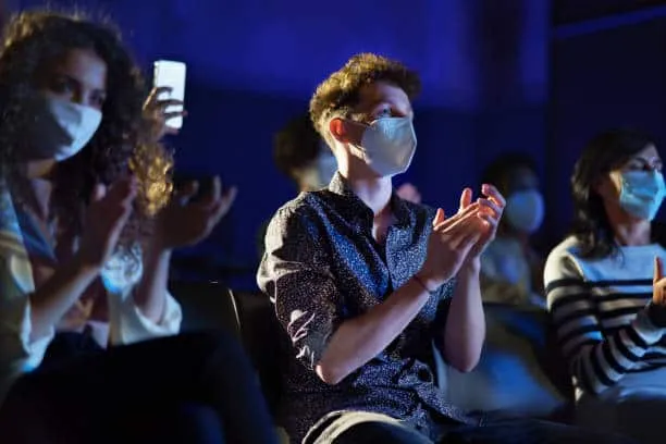 People wearing face masks attending a concert or event, clapping and enjoying entertainment in a dark indoor setting, emphasi