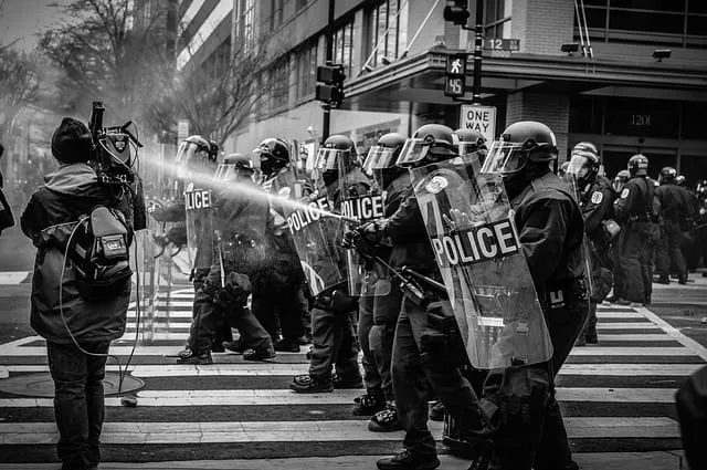 Police officers in riot gear with shields during a protest on a city street, black and white image capturing civil unrest and