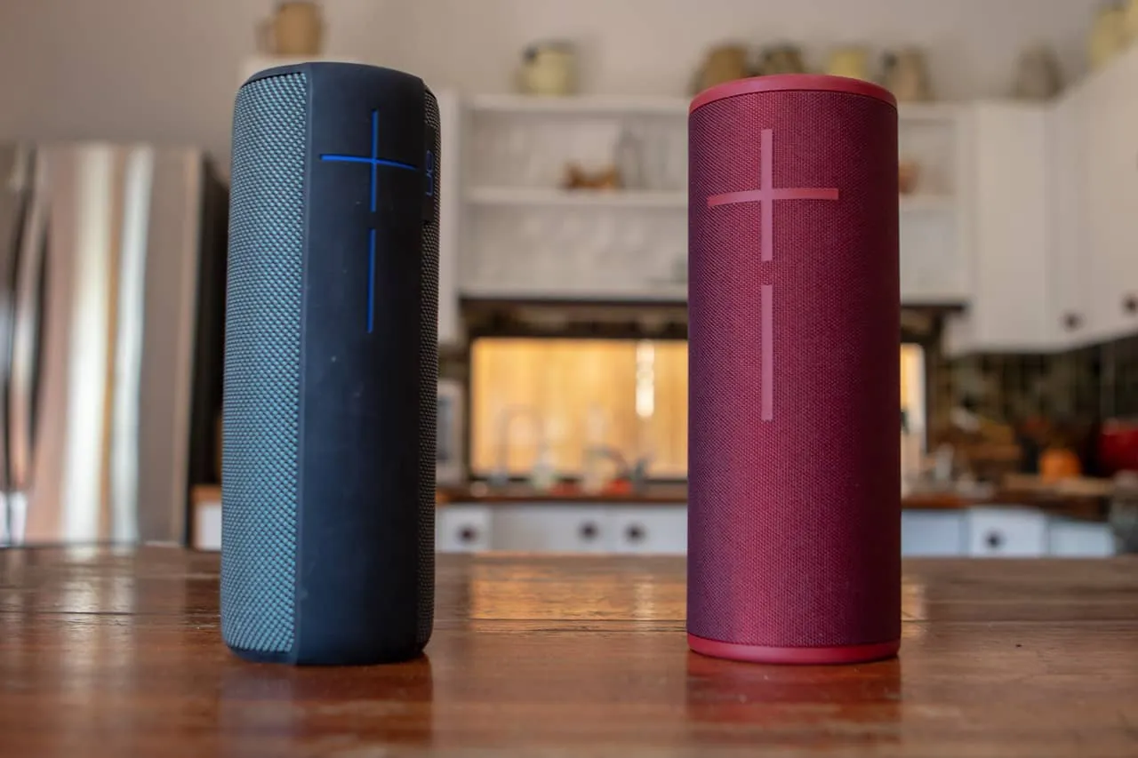 Wireless portable Bluetooth speakers with volume controls, black and red, on a wooden kitchen table.