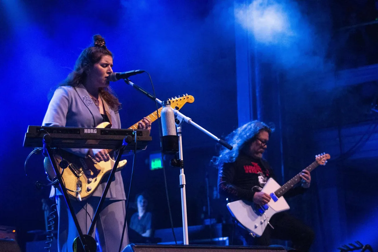 Performing musicians on stage with guitars and keyboard during a live concert, illuminated by blue stage lighting, showcasing