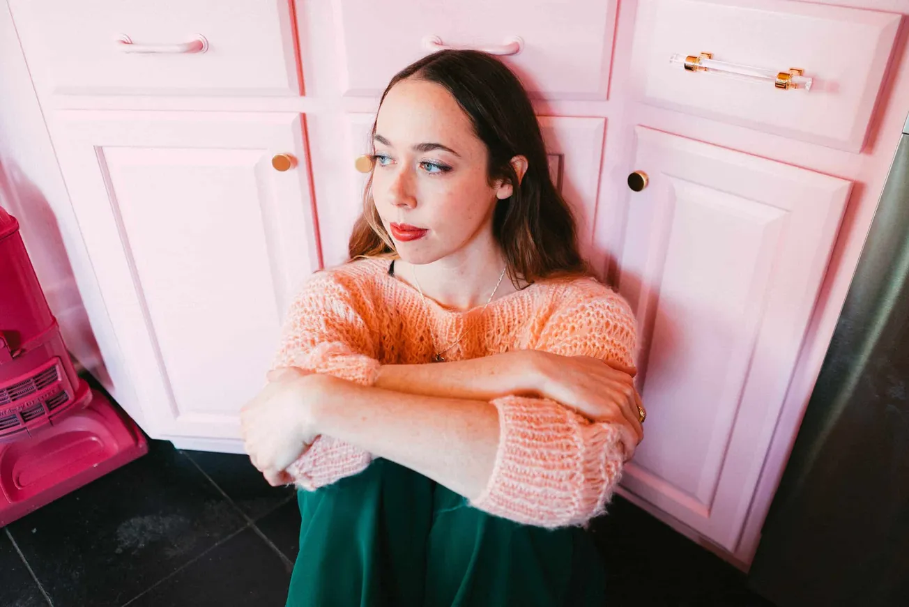 Soft pink kitchen cabinets with gold accents create a stylish backdrop for a young woman in a cozy peach sweater seated on th