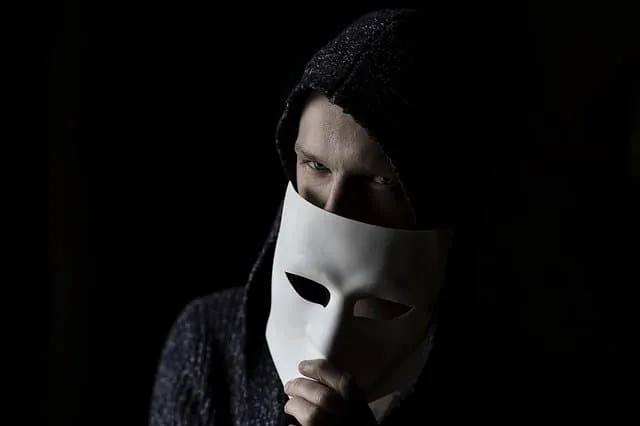 Mysterious person wearing a black hoodie and holding a white mask in a dark setting, symbolizing anonymity or secret identity