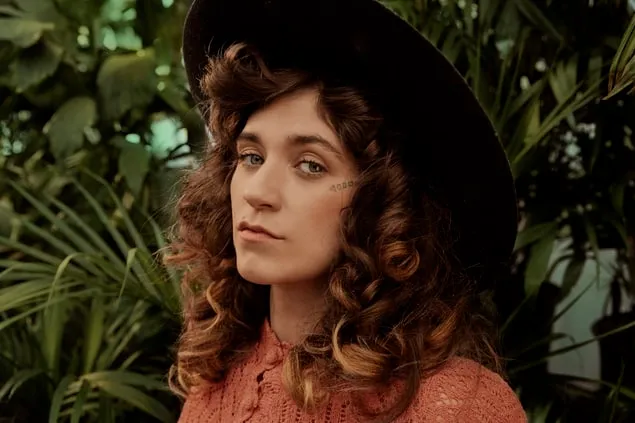Fashion portrait of a young woman with curly hair wearing a wide-brimmed hat and a pink textured top, surrounded by lush gree