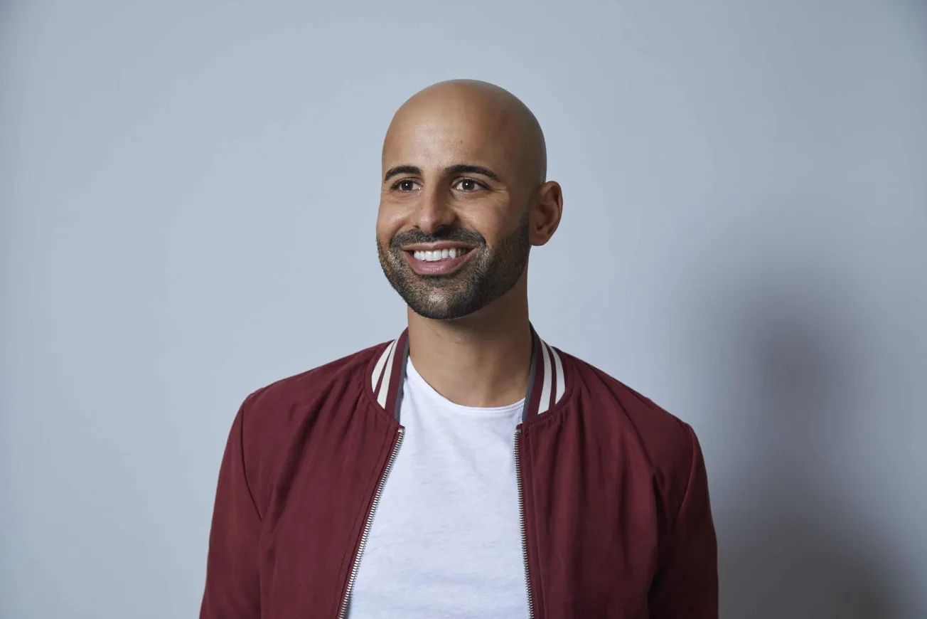 Bald man with beard smiling wearing a maroon jacket and white t-shirt against a plain light gray background.