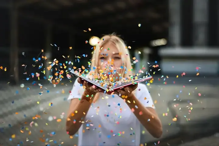 Bright young woman celebrating with colorful confetti and an open book outdoors, vibrant and joyful, illustrating excitement,