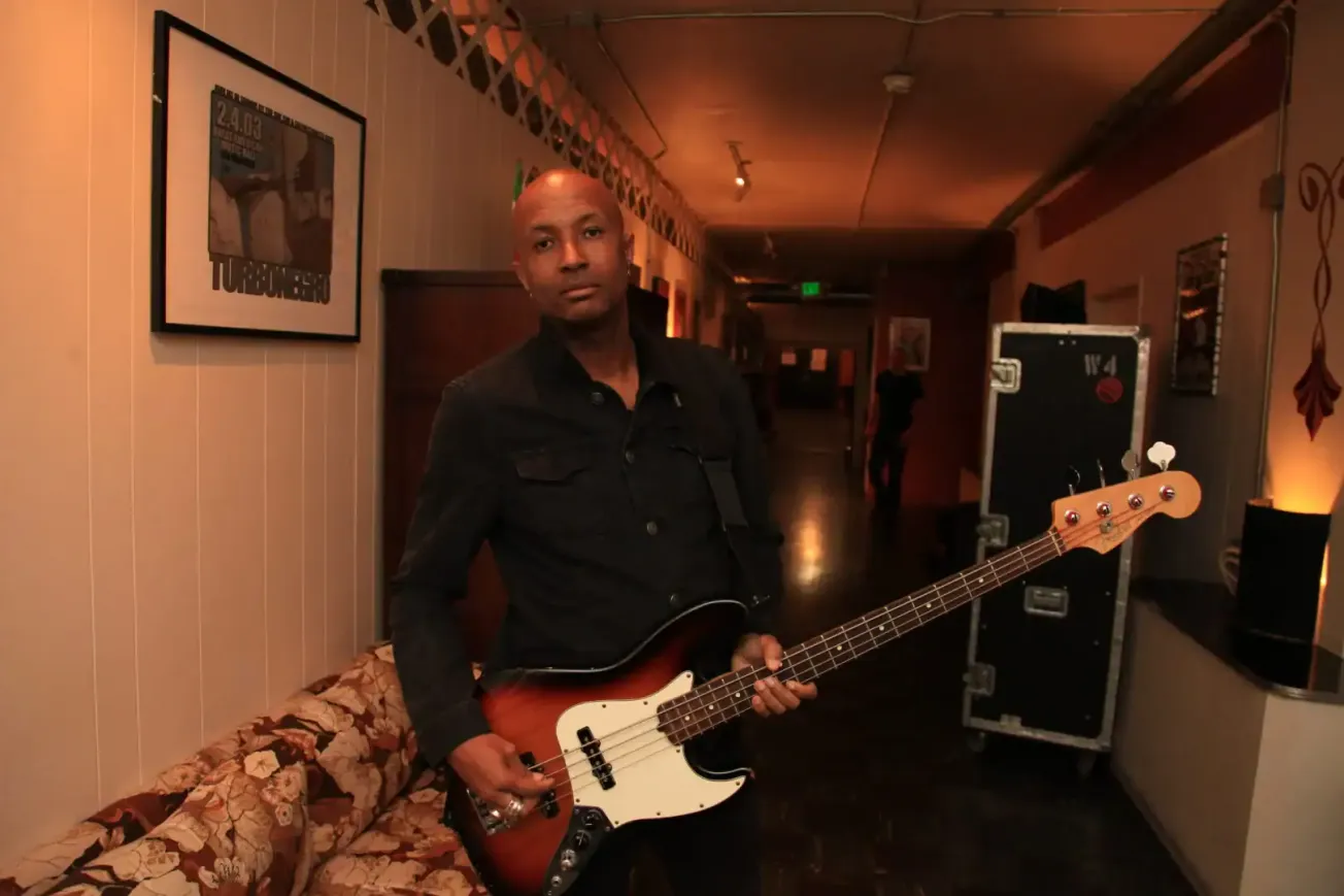 A musician with a bass guitar standing in a warmly lit, cozy venue, ready to perform or practice, surrounded by framed concer