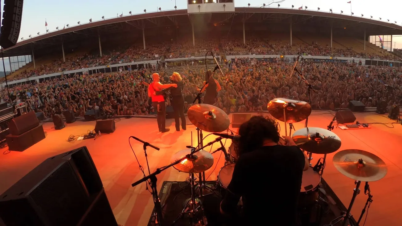 Vibrant live music performance at an outdoor concert with a large, enthusiastic audience enjoying the show. The stage feature