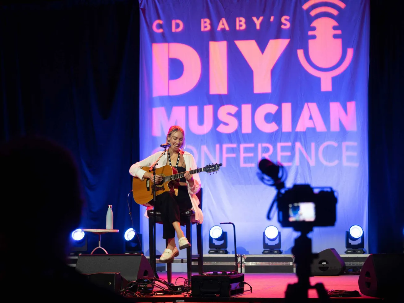 Girl performing live on stage playing guitar and singing at CD Baby's DIY Musician Conference, with professional camera recor