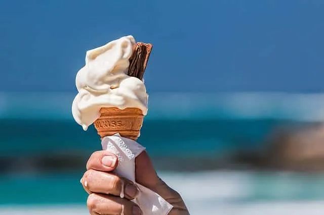 A hand holding a melting ice cream cone with chocolate wafer stick against a blue sky and ocean background.