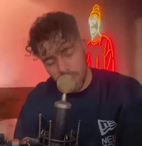 A young man with curly hair singing into a microphone in a cozy, dimly lit room with neon artwork in the background, capturin
