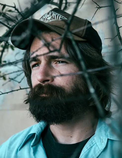 Man with a beard wearing a cap and a turquoise shirt, looking thoughtfully through tree branches.
