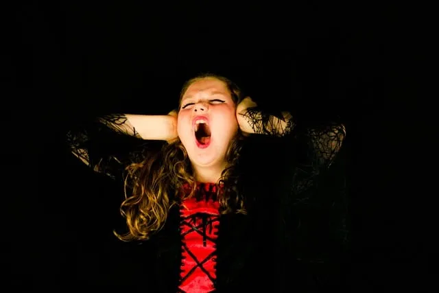 A young girl with curly hair covering her ears, screaming or shouting with a distressed expression against a black background