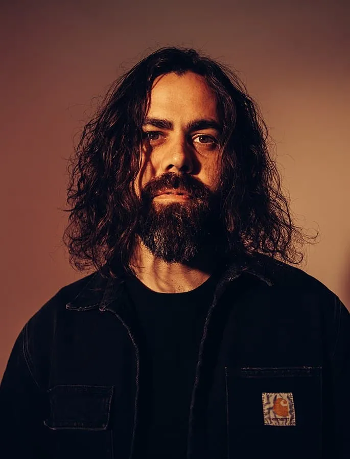 Portrait of a man with long curly hair and a beard, wearing a black denim jacket, against a warm-toned background, highlighti