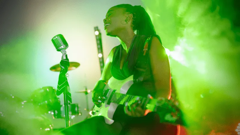 Women musician performing live with guitar and microphone during concert, vibrant green stage lighting, highlighting the musi