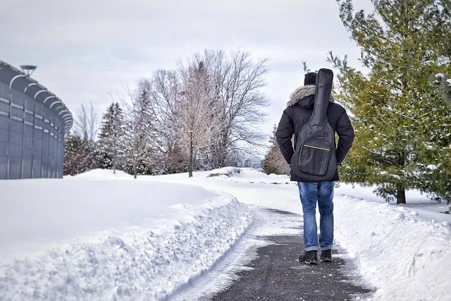 A person walking on a snowy path with a guitar case on their back, winter landscape with snow-covered trees and buildings in