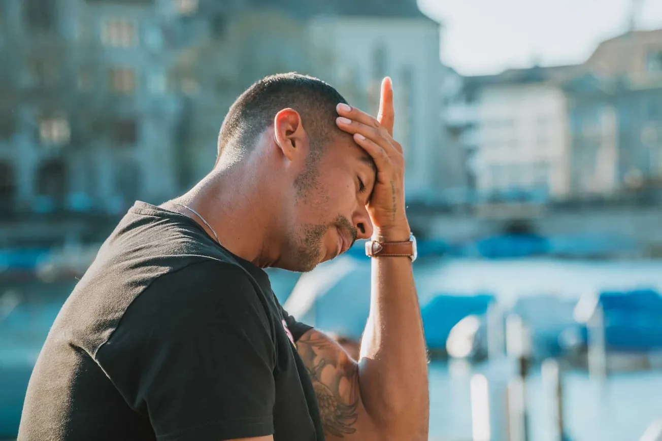 A man with tattoos in distress holding his forehead near a harbor with boats, illustrating stress or worry in an urban outdoo