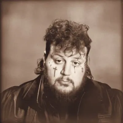 A man with tattoos on his face, curly hair, and a leather jacket, posing against a plain background.