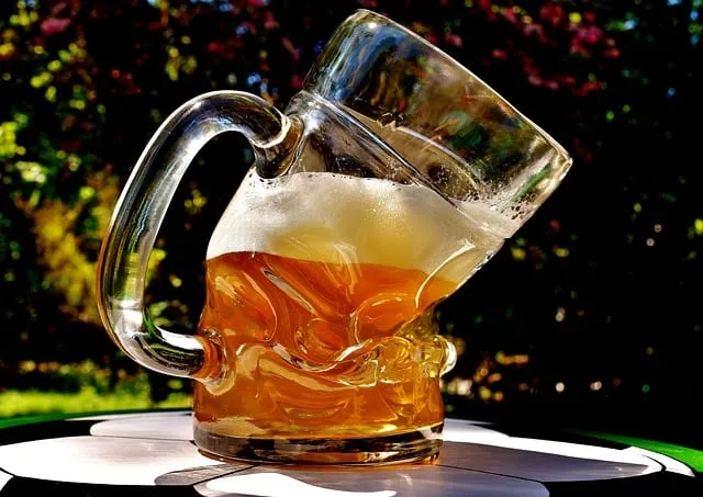 A melting glass mug of beer with foam on the rim, tilted at an angle outdoors with a blurred natural background.