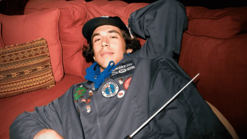 Relaxing young man lying on sofa with musical conductor baton, casual attire with patches, cozy home setting, leisure and mus