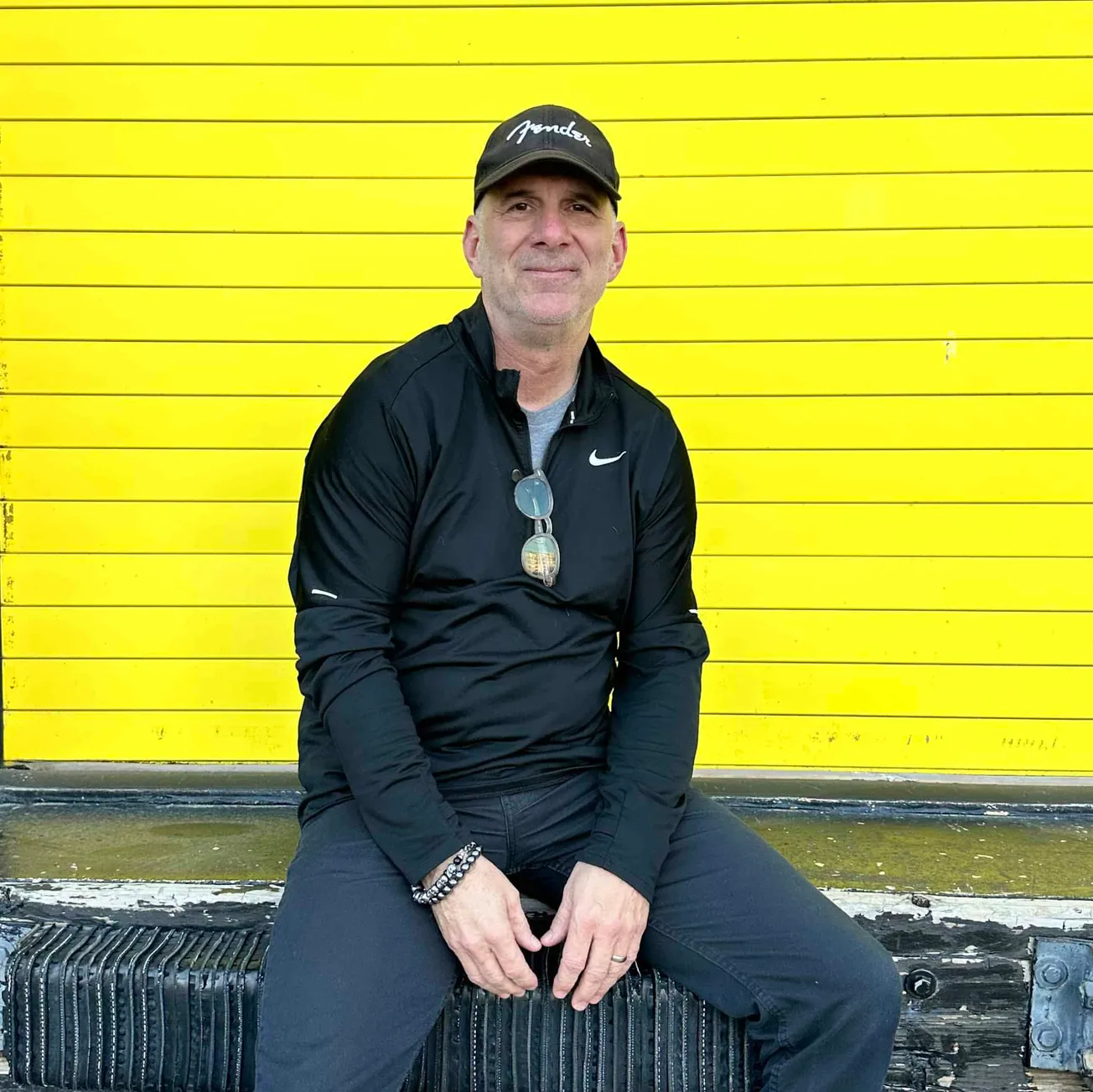 Colorful yellow background wall with a man sitting on a black outdoor bench, dressed in black Nike athletic wear, wearing sun