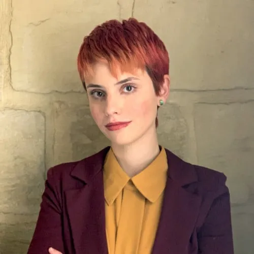 Bright red short haircut of a confident woman in a mustard blouse and blazer against a stone wall, representing modern music