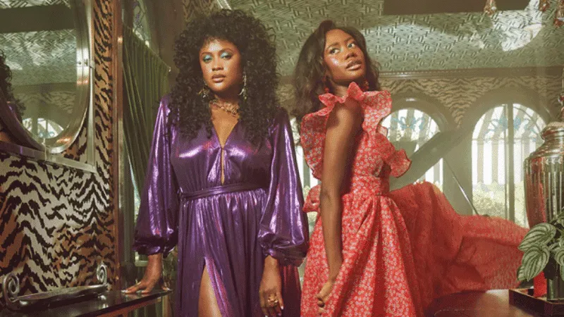 Fashion-forward women wearing glamorous dresses in a stylish, vibrant indoor setting with patterned decor and lush greenery,